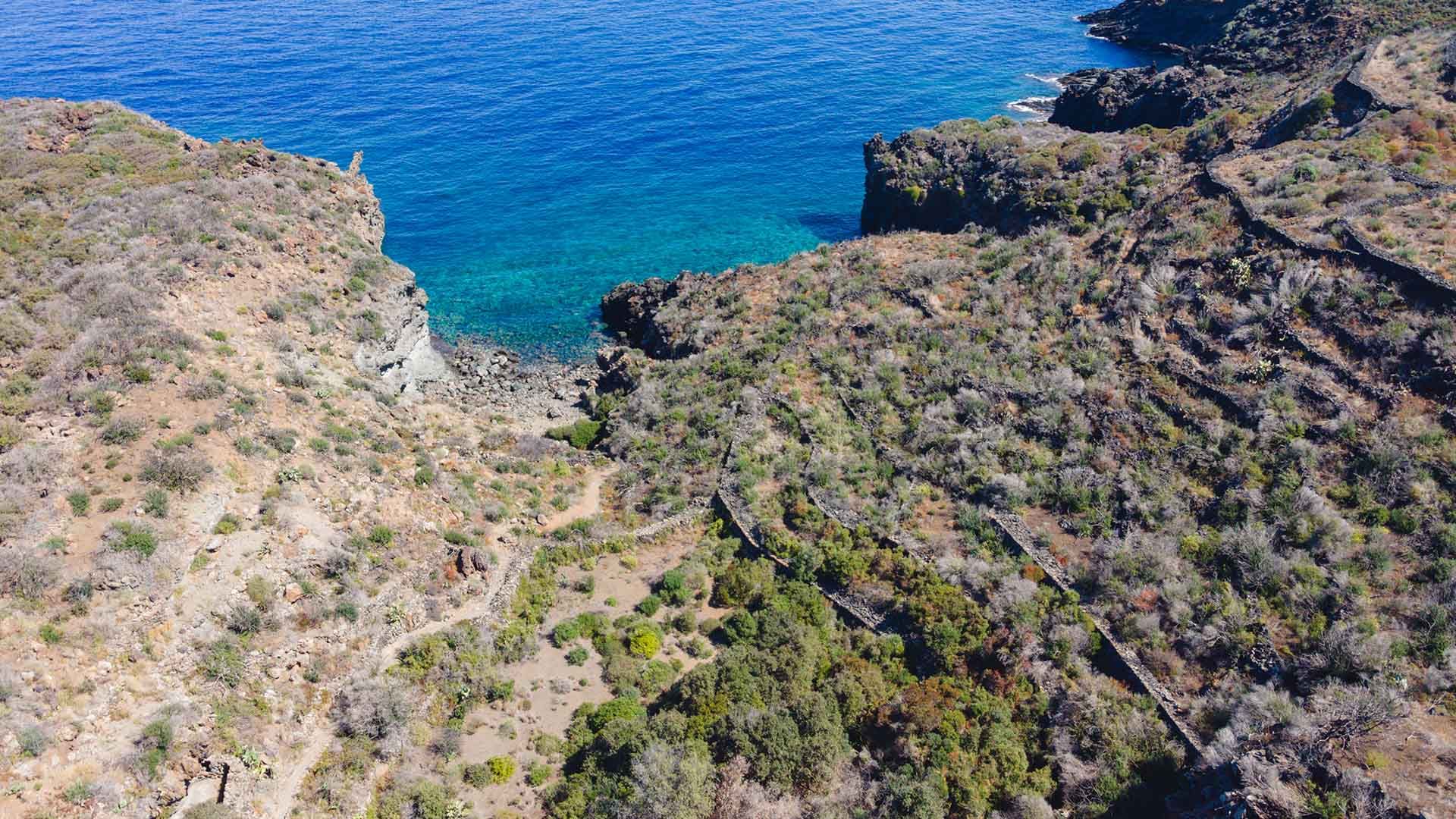 Veduta aerea di una piccola insenatura circondata da colline aride e terrazzamenti in pietra, con mare limpido e vegetazione sparsa.