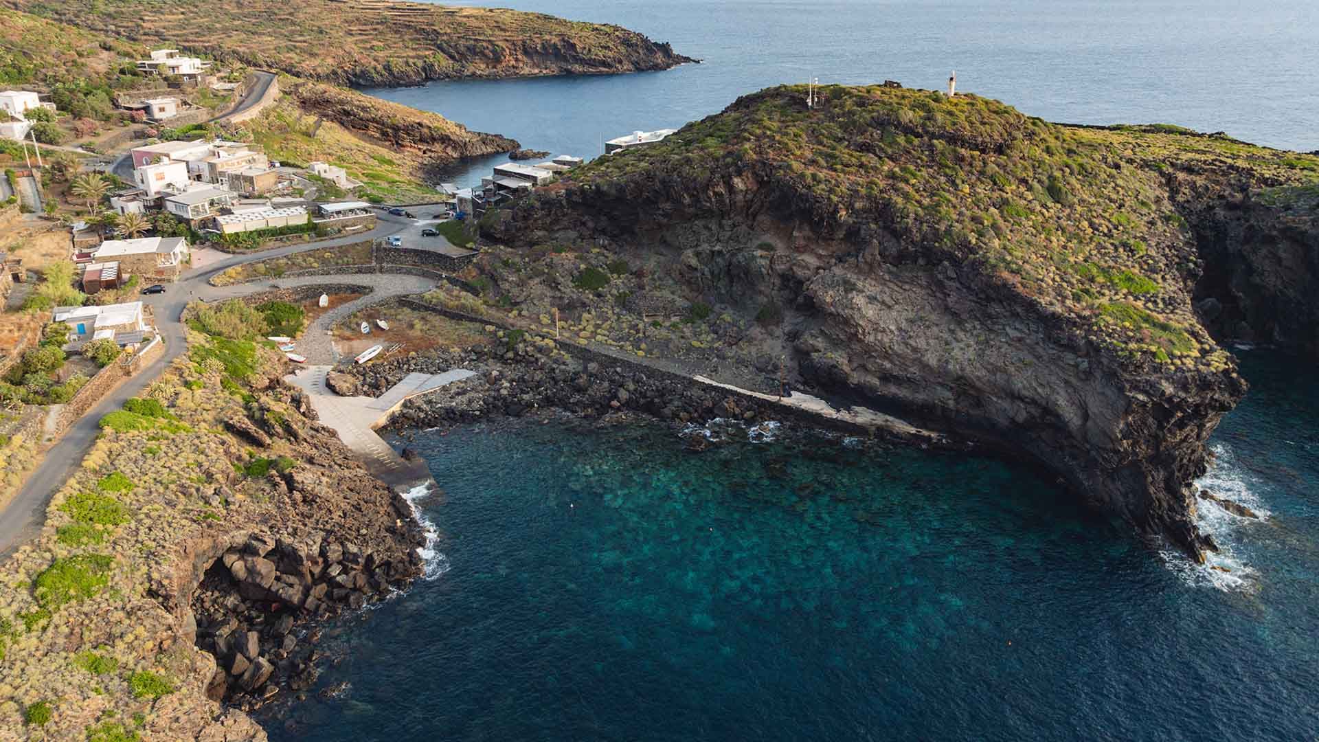 Veduta panoramica di una cala rocciosa con case sparse sul promontorio, un piccolo porticciolo e acque turchesi, circondata da scogliere verdi.