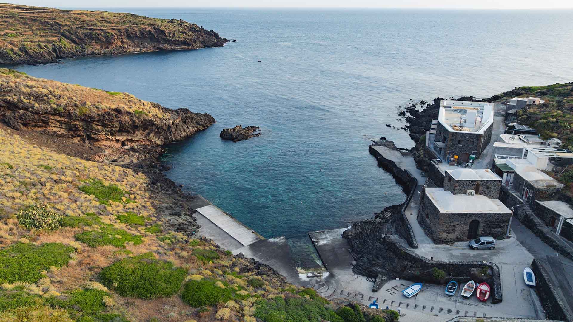 Veduta dall’alto di una piccola cala protetta da scogliere rocciose e vegetazione mediterranea, con edifici in pietra lavica a ridosso del mare e alcune barche tirate a secco.