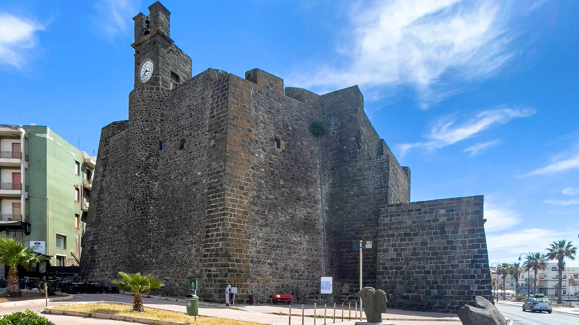 Massiccio castello in pietra con torre dell’orologio, situato in un contesto urbano con edifici sullo sfondo e cielo sereno.