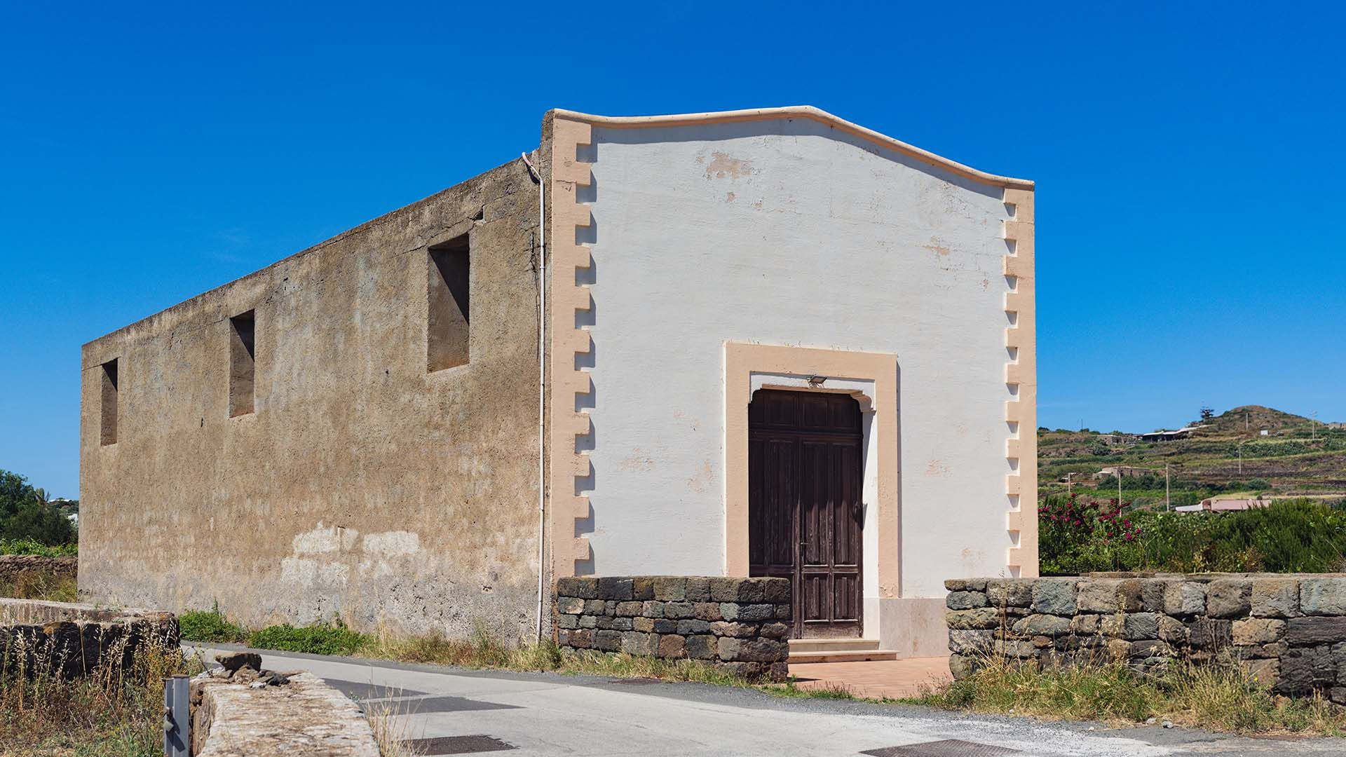 Chiesa rurale semplice con facciata intonacata e ingresso in legno scuro, circondata da paesaggio agricolo e cielo limpido.