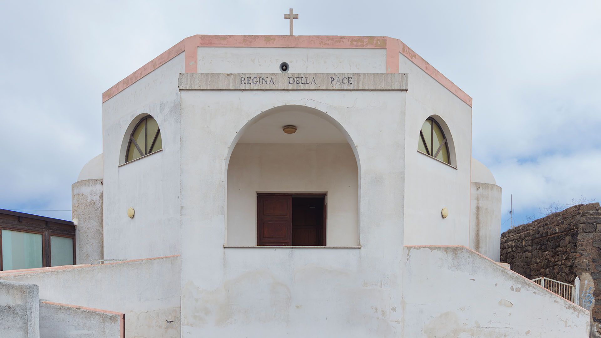 Facciata della Chiesa della Madonna della Pace a Pantelleria, edificio moderno a tinte chiare con croce in cima, archi e finestre semicircolari. Sulla facciata è incisa la scritta “Regina della Pace”. Il cielo è coperto da nuvole chiare.