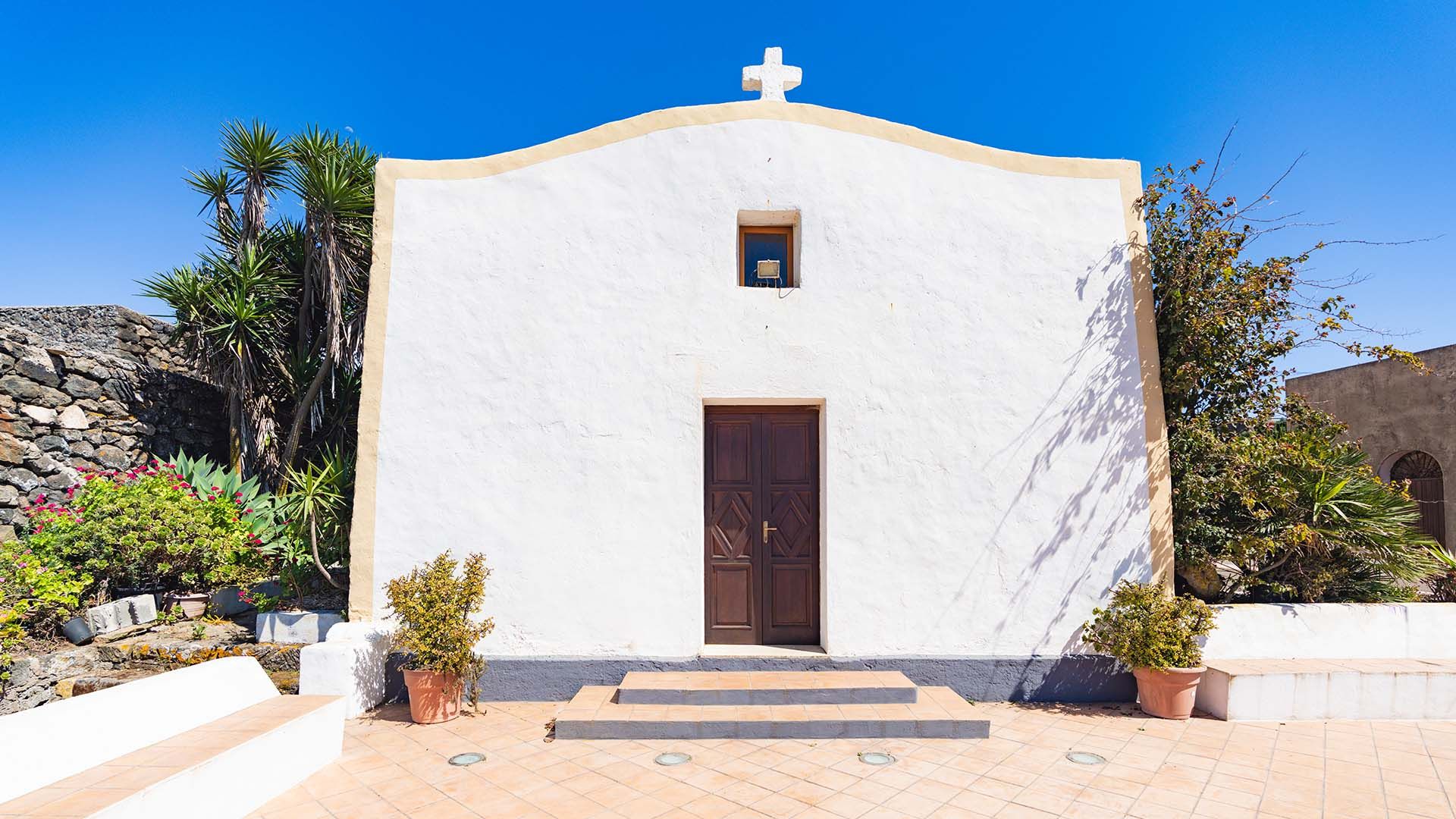 Facciata della Chiesa della Madonna del Rosario a Pantelleria, con pareti bianche e profilo curvo, croce bianca in cima e porta in legno scuro al centro. Ai lati, vasi con piante e vegetazione mediterranea, sotto un cielo azzurro intenso.