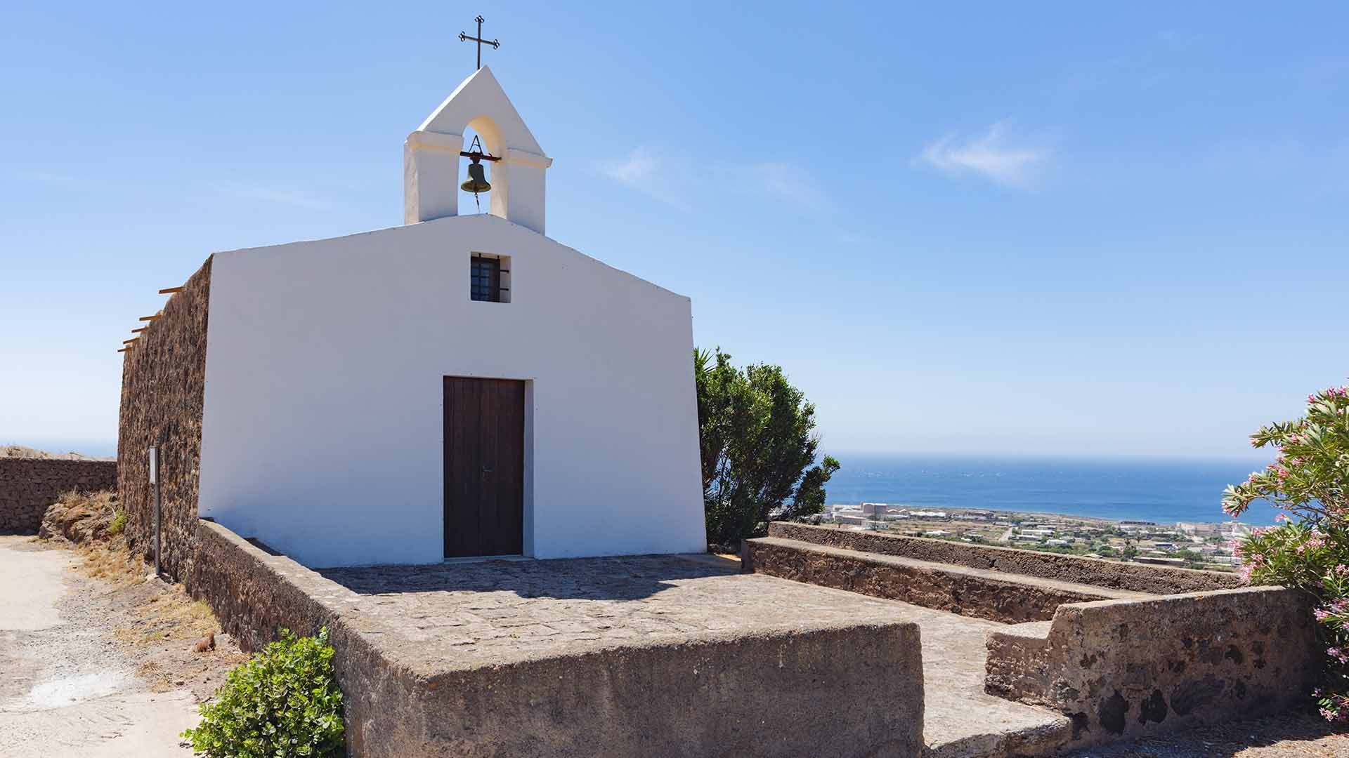 Piccola chiesa bianca con campanile a vela, situata in posizione panoramica su un'altura con vista sul mare e sul paesaggio urbano sottostante.