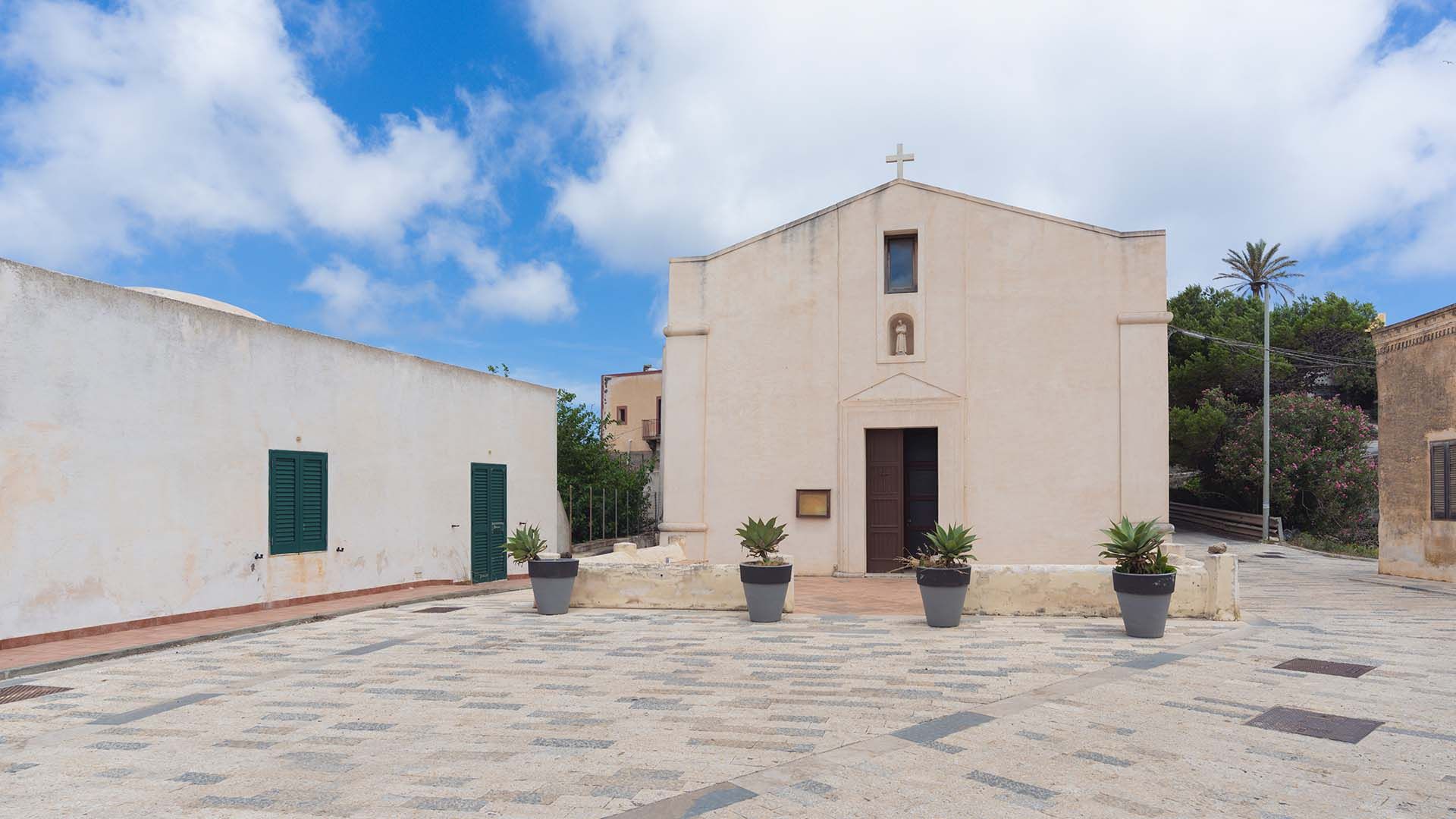 Facciata semplice color crema della Chiesa di San Francesco, con piccolo campanile e croce in cima, situata in una piazza lastricata con vasi decorativi e cielo parzialmente nuvoloso.