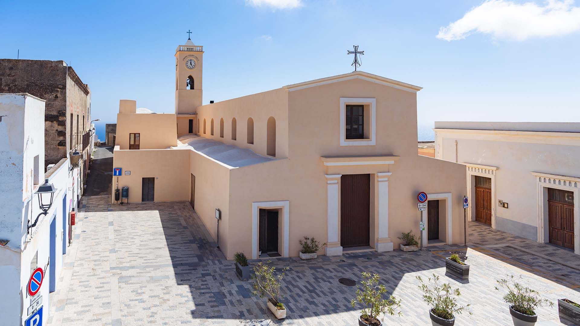 Chiesa dal colore chiaro con campanile a torre e orologio, situata in una stretta via lastricata, con vista sul mare sullo sfondo e cielo limpido.