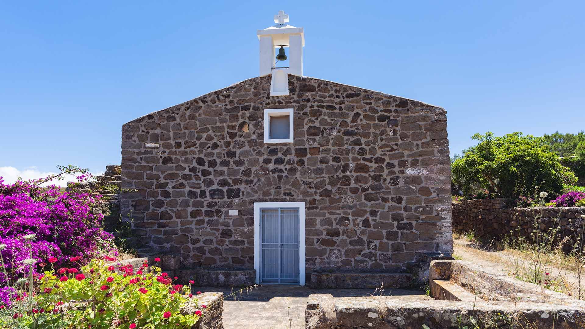 Piccola chiesa in pietra scura con campanile a vela e croce bianca, immersa nella vegetazione e circondata da fiori colorati e muri in pietra.