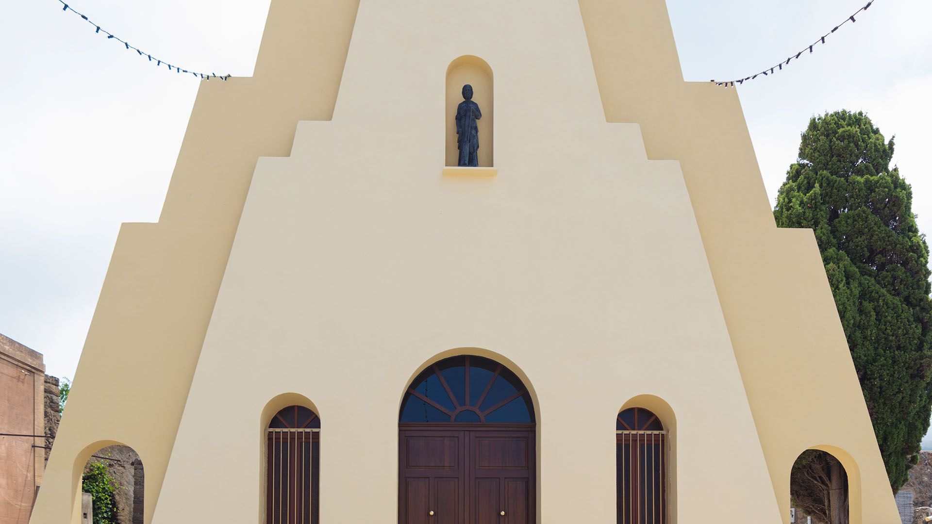 Chiesa con facciata triangolare a gradoni color sabbia e grande portone in legno, con una statua scura in una nicchia centrale e decorazioni luminose sopra.