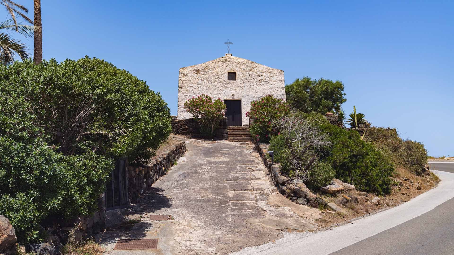 Chiesa rurale in pietra chiara su una collinetta, raggiungibile tramite una rampa in cemento tra cespugli e palme, con semplice croce in ferro sulla sommità.
