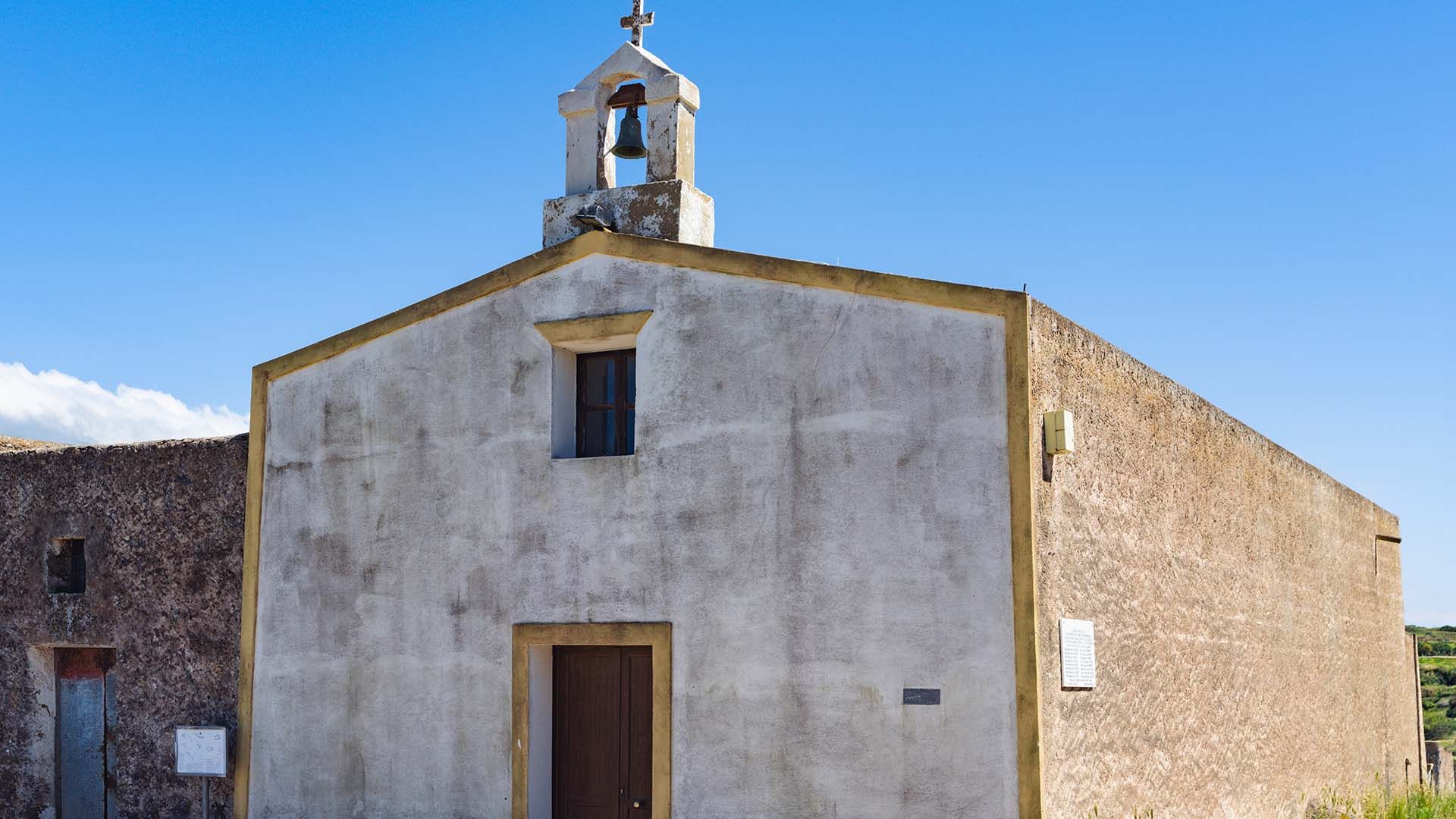Chiesa semplice con facciata intonacata e campanile a vela con campana, porta in legno e finestrella centrale, sotto un cielo limpido e blu.