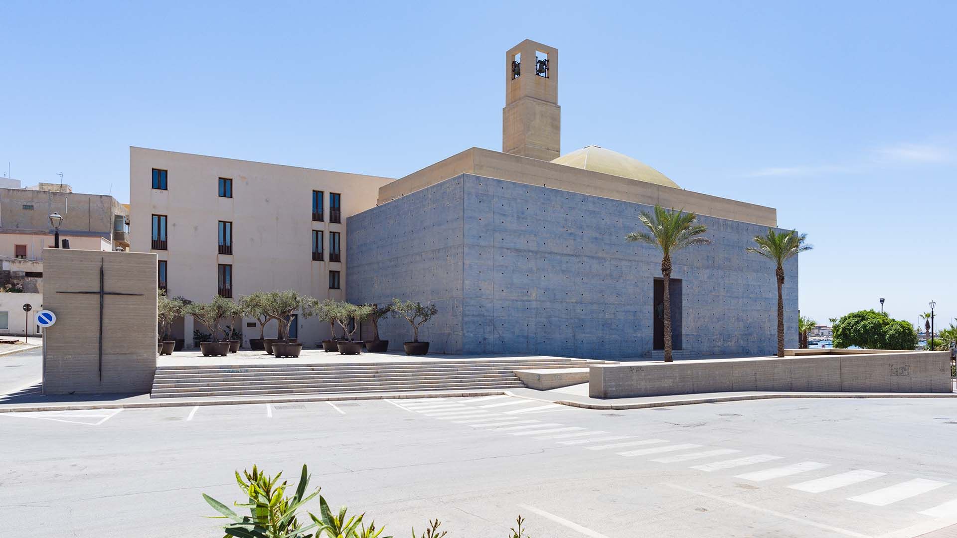 Veduta della Chiesa del SS. Salvatore a Pantelleria, edificio moderno in cemento con pareti lisce e squadrate, cupola gialla e torre campanaria con doppia campana. Davanti, una piazza con gradinate, palme e ulivi in grandi vasi, sotto un cielo limpido.
