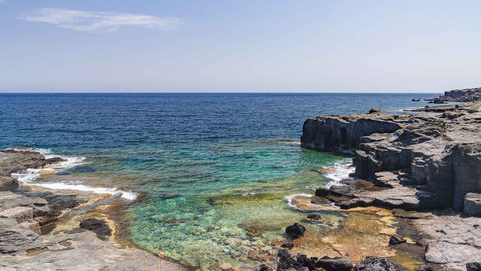Caletta del Bue Marino a Pantelleria, caratterizzata da scogliere di pietra lavica e fondali rocciosi con acque trasparenti che sfumano dal verde al blu. In lontananza, l’orizzonte marino si fonde con un cielo sereno.