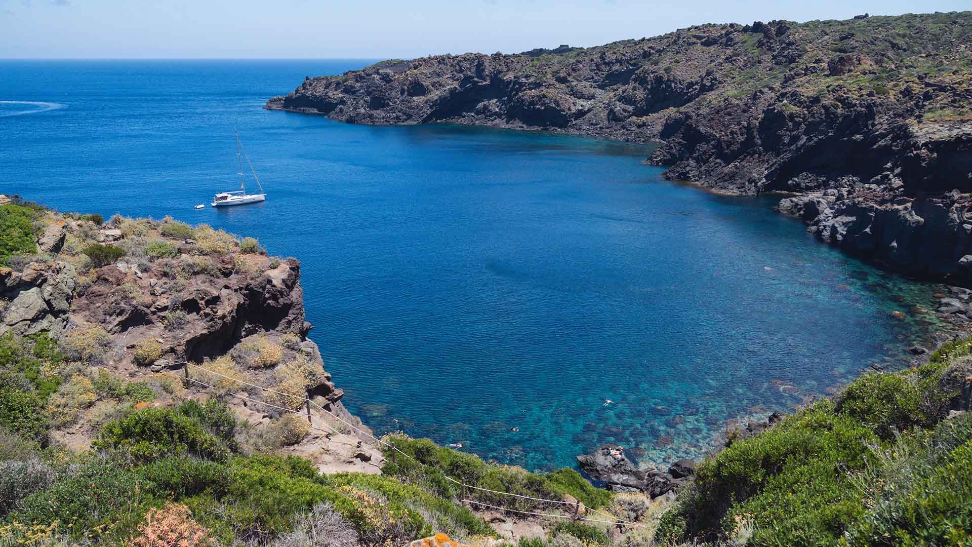 Veduta panoramica della Cala Cinque Denti a Pantelleria, con acque limpide e blu circondate da alte scogliere rocciose. In primo piano, vegetazione mediterranea e un sentiero che scende verso il mare, dove è ancorato un catamarano bianco.