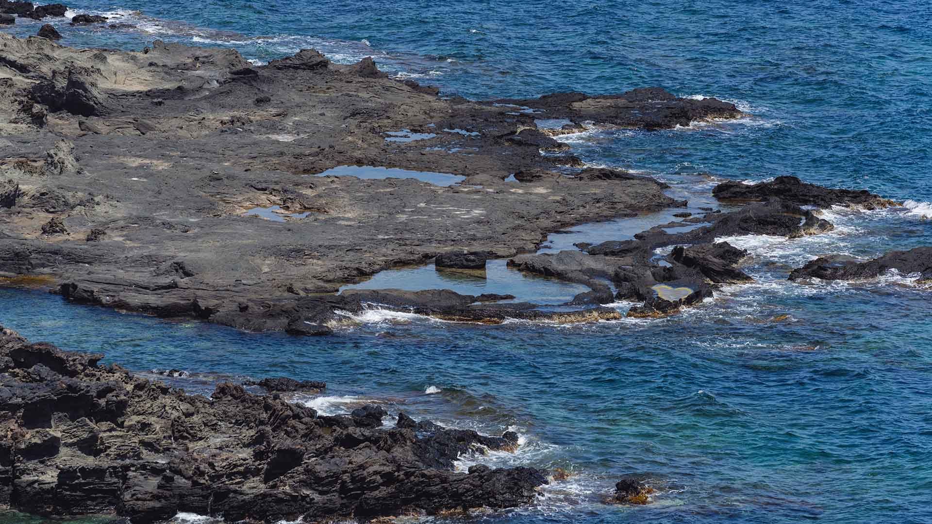 Costa lavica con pozze scure d’acqua e scogli affacciati su un mare blu intenso, con leggere increspature delle onde.