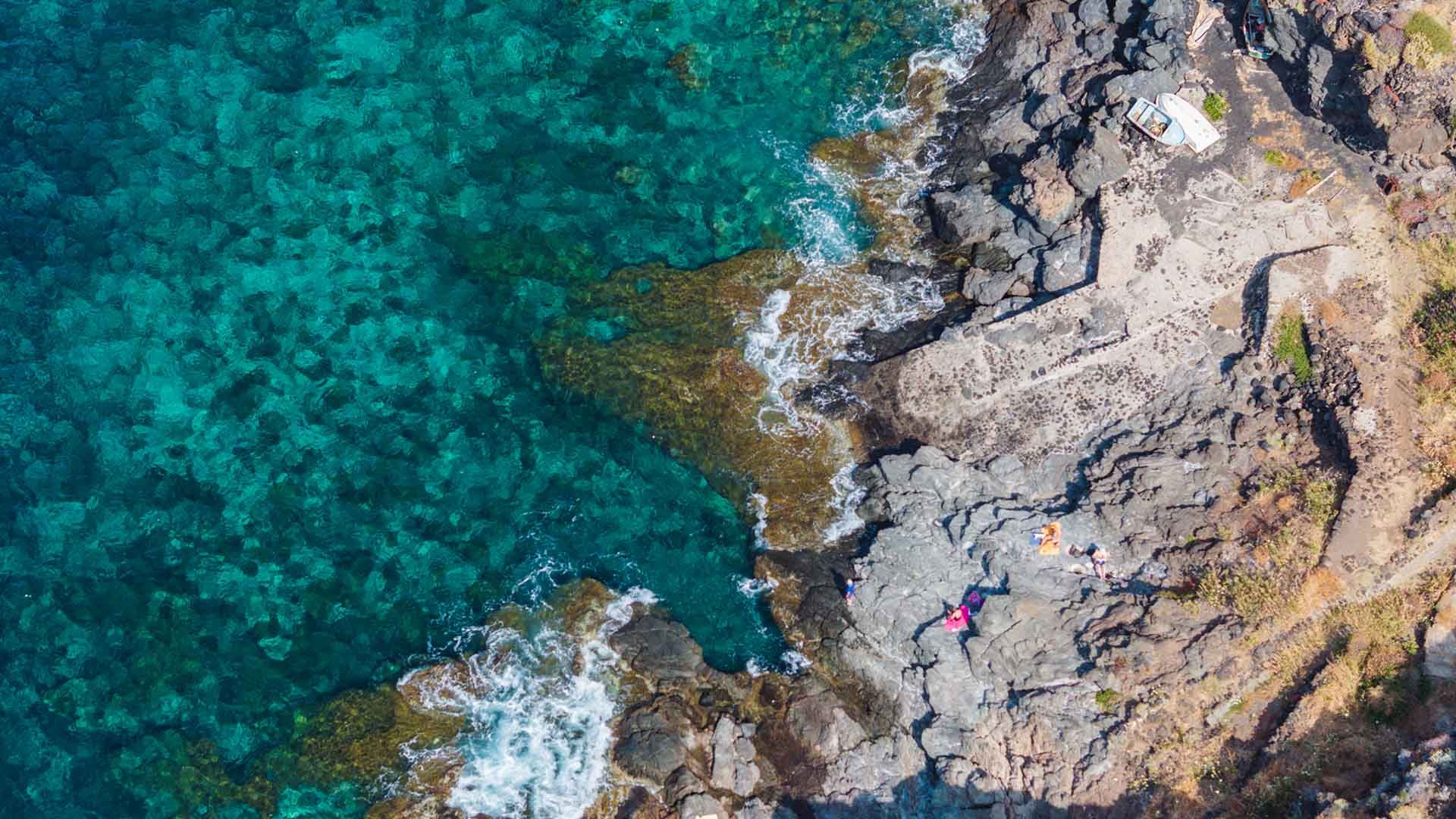 Ripresa aerea di una costa rocciosa con mare cristallino dai riflessi turchesi. Sulla destra si nota una piccola piattaforma in cemento con barca ormeggiata e persone distese a prendere il sole sugli scogli scuri. L’acqua trasparente lascia intravedere il