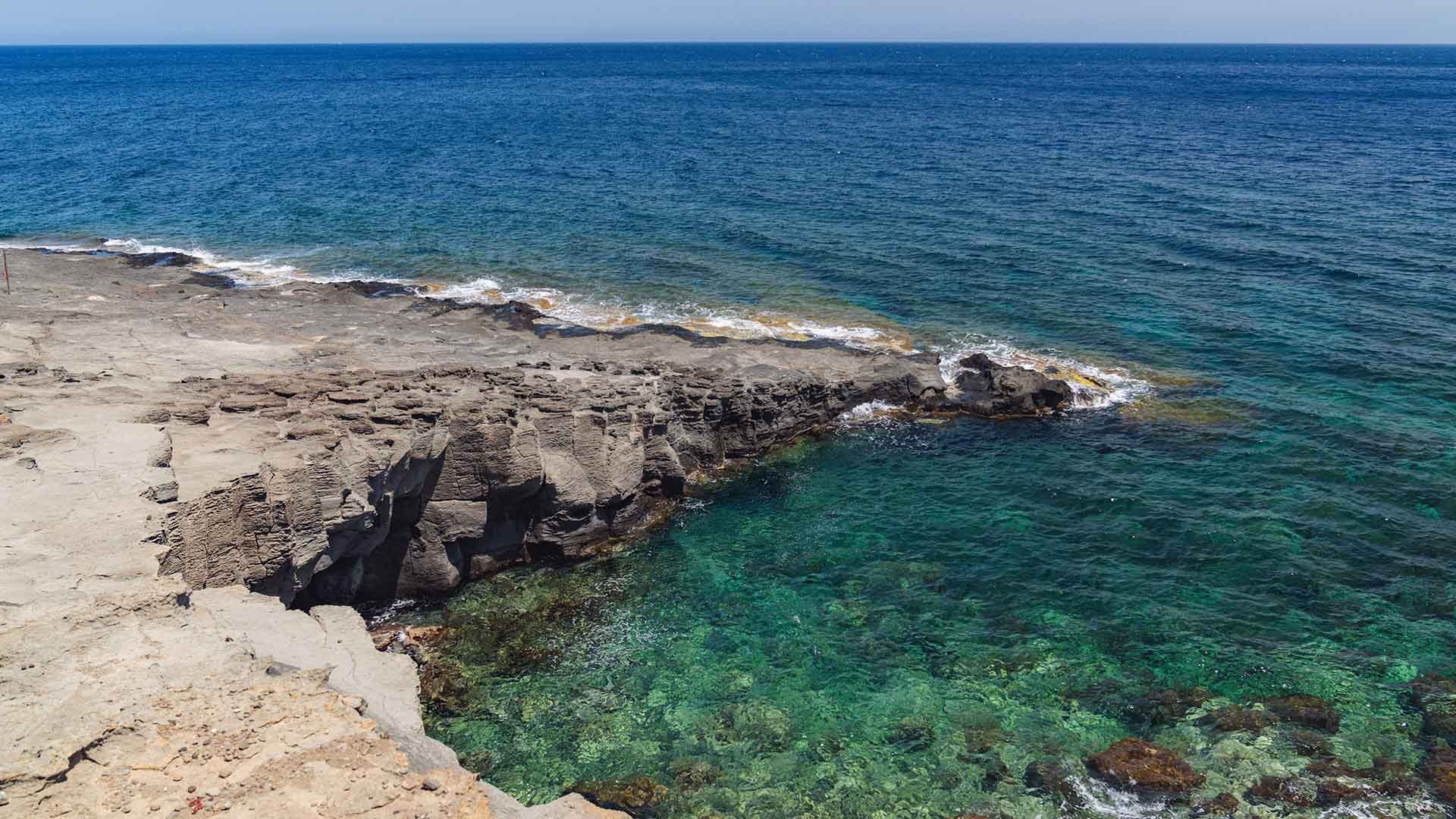 Scogliera vulcanica scolpita dal tempo che si protende verso un mare cristallino. In primo piano, la roccia grigia contrasta con il verde smeraldo dell’acqua bassa, mentre sullo sfondo l’orizzonte si apre su un mare blu profondo.