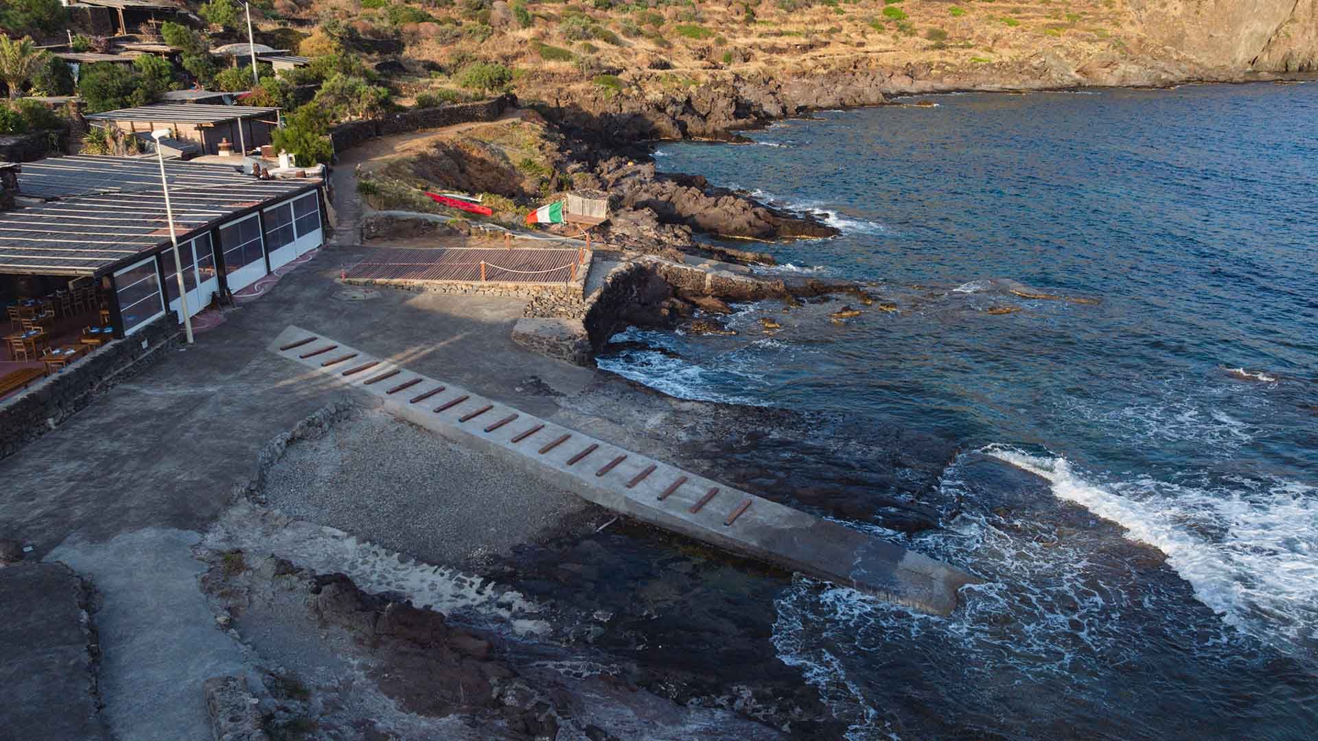 Scalo a mare nella località di Scauri a Pantelleria, con una passerella in cemento che si estende sugli scogli fino al mare. A sinistra, strutture coperte con tavoli e sedie; sullo sfondo, una costa rocciosa e vegetazione mediterranea.