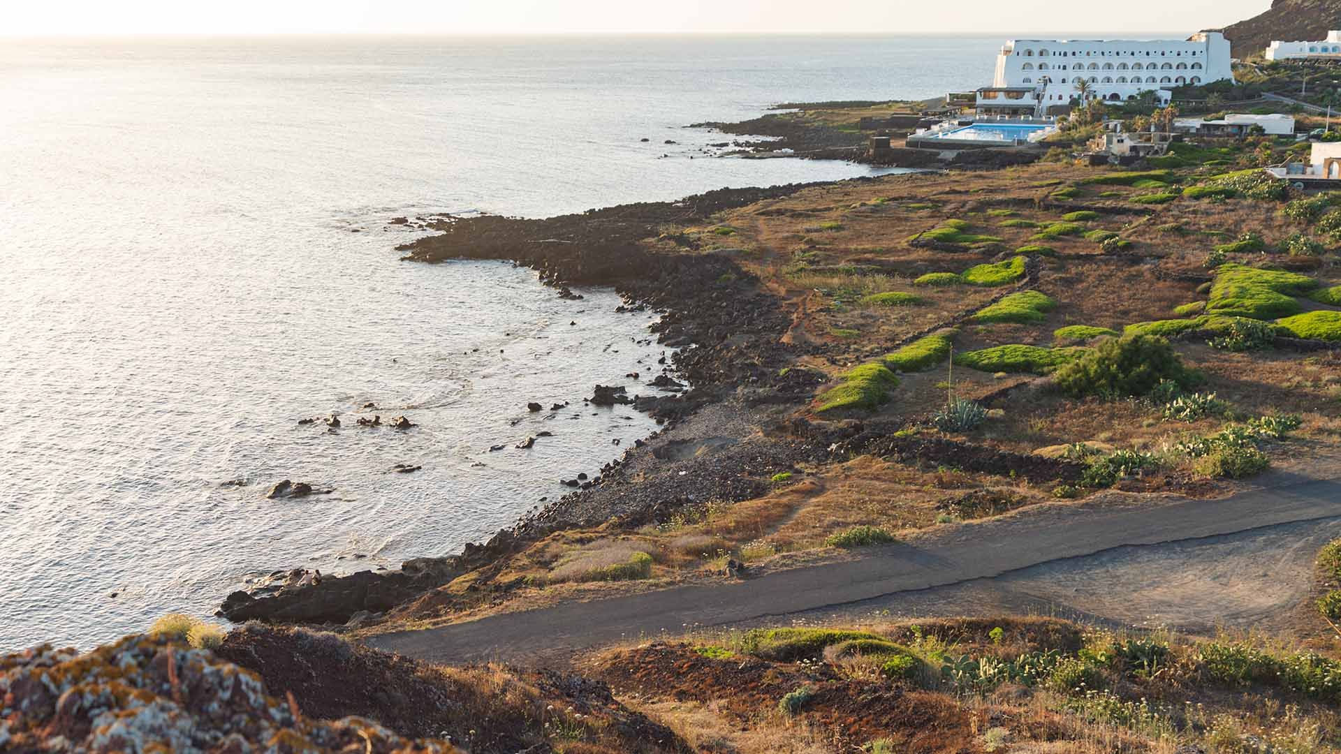 Vista costiera al tramonto con una scogliera lavica che degrada dolcemente verso il mare calmo. In primo piano un paesaggio arido punteggiato da cespugli verdi, mentre sullo sfondo si distingue un grande edificio bianco con piscina affacciato sul mare.