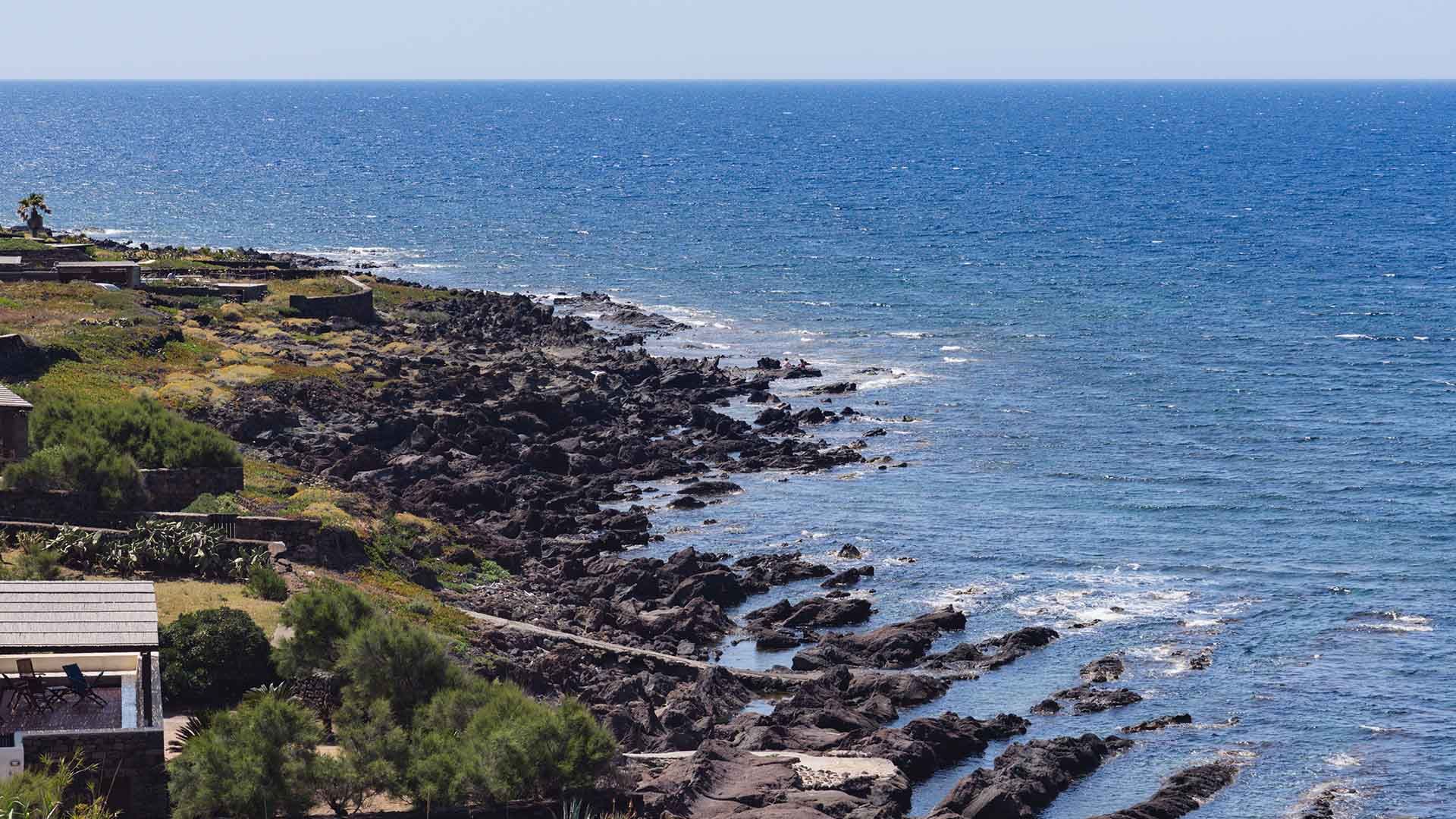 Tratto costiero frastagliato e roccioso, dove la vegetazione mediterranea incontra le scure formazioni laviche che scendono verso il mare. In basso a sinistra si intravedono delle strutture con terrazze vista mare, mentre lo sguardo si perde tra le onde e