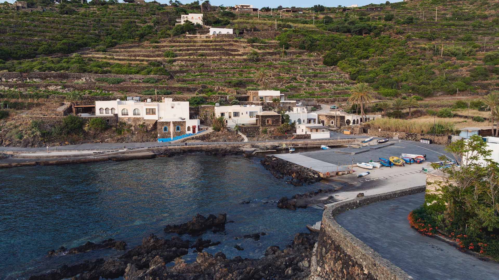 Veduta del piccolo borgo marinaro di Gadir a Pantelleria, con case bianche affacciate su un porticciolo circondato da terrazze coltivate e muretti a secco. Alcune barche sono tirate a secco sulla banchina, mentre il mare limpido lambisce la costa rocciosa