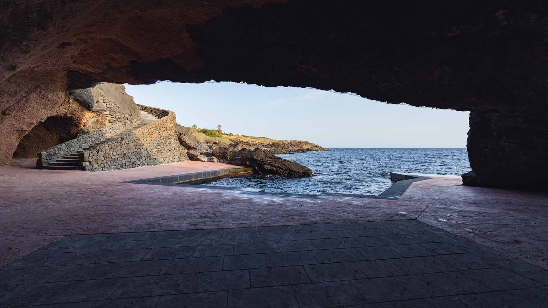 Vista dall’interno di una grotta sul mare con pavimento e gradinate in pietra lavorata, che si apre verso una piccola baia con acque limpide. L’accesso al mare è incorniciato da archi naturali nella roccia, mentre la luce del tramonto illumina dolcemente 