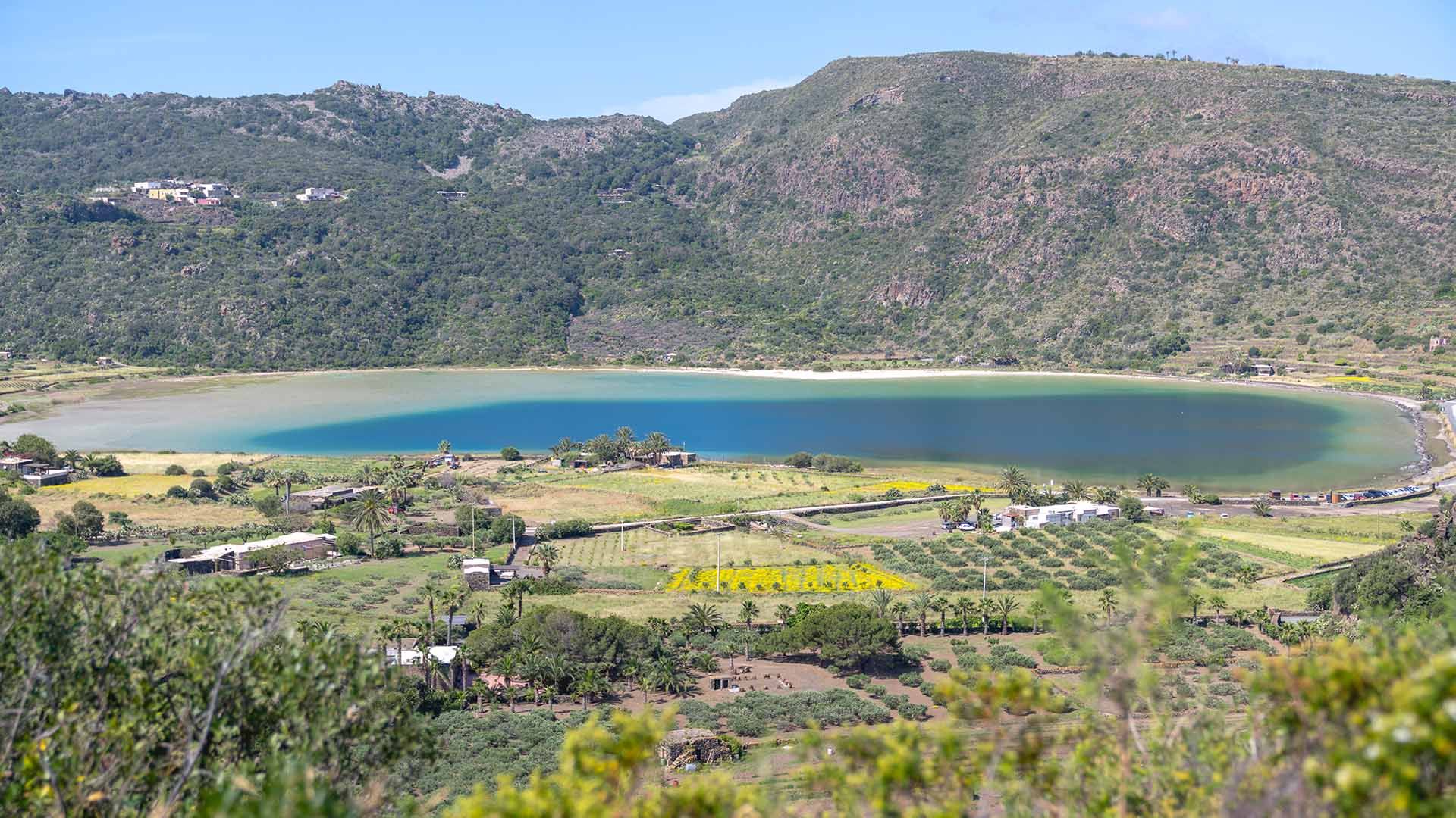 Veduta panoramica del Lago di Venere, uno specchio d’acqua turchese di origine vulcanica situato nel cratere di un antico vulcano, circondato da colline verdeggianti e campi coltivati. In primo piano si notano palme, ulivi e costruzioni rurali, mentre l’a