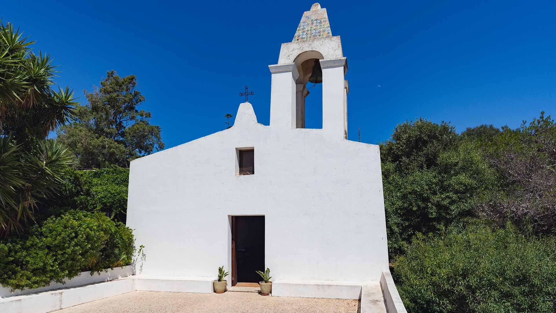 Facciata della Chiesa della Madonna delle Grazie a Pantelleria, semplice e intonacata di bianco, con campanile a vela decorato da maioliche colorate. Due piccole piante in vaso ai lati della porta d’ingresso e vegetazione rigogliosa tutt’intorno sotto un 