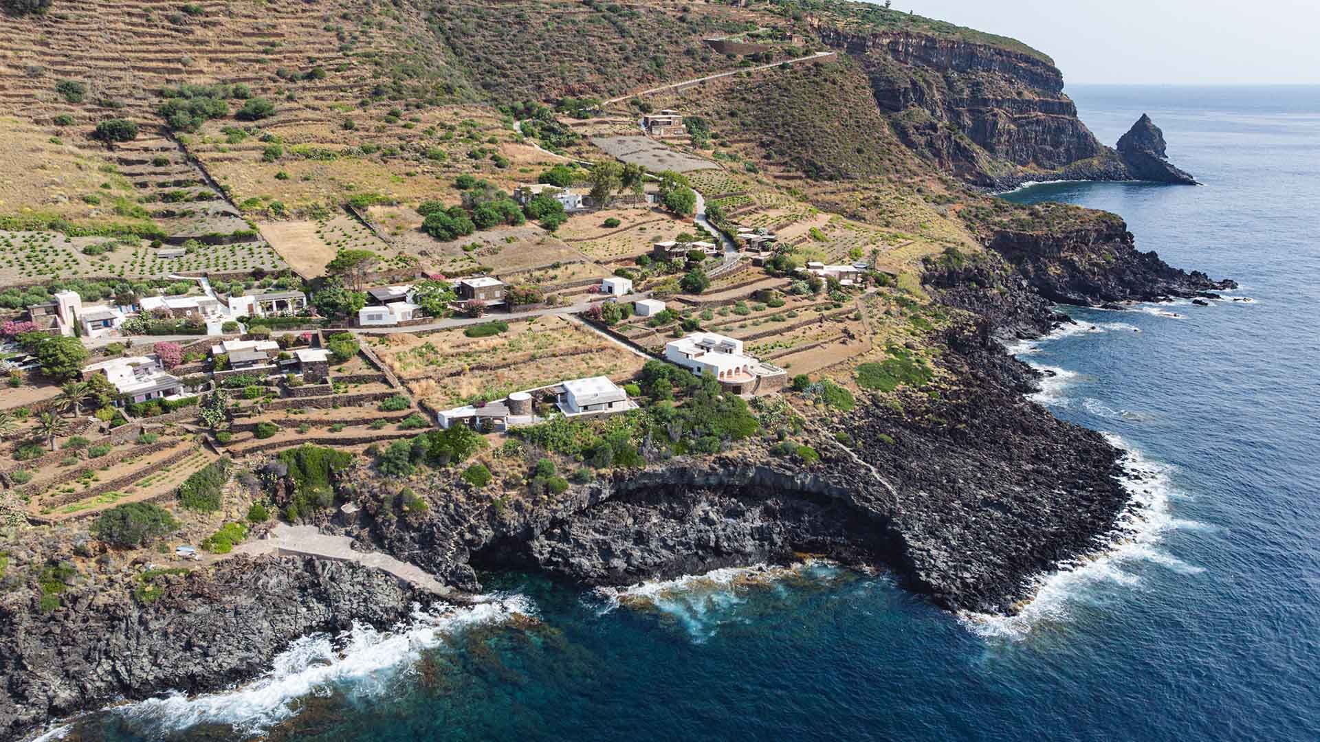 Vista aerea della contrada di Martingana a Pantelleria, con case bianche sparse lungo una scogliera vulcanica scura. I terrazzamenti coltivati coprono i pendii della collina, mentre il mare azzurro e profondo lambisce le rocce sottostanti. Sullo sfondo, u