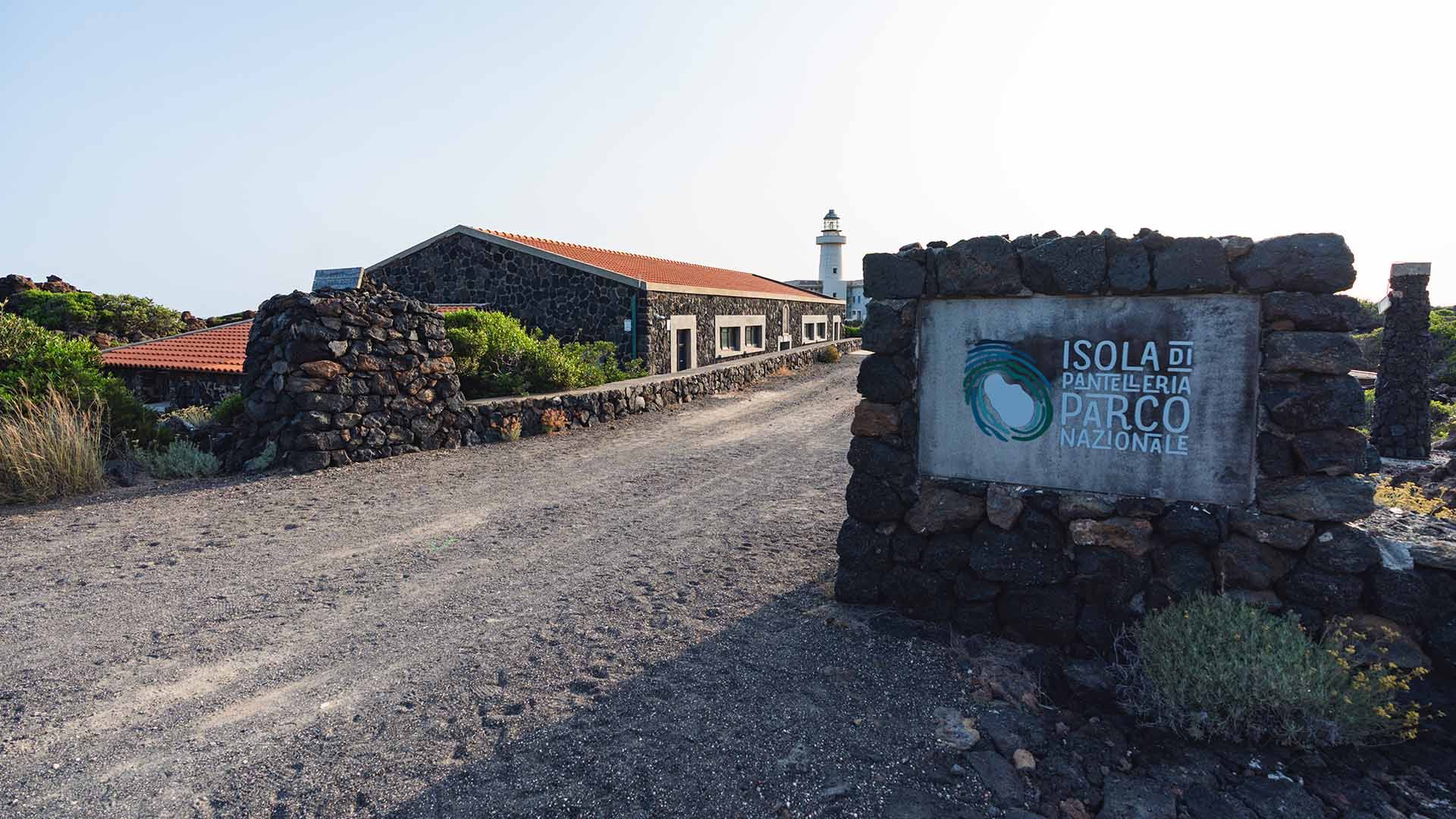 Ingresso sterrato con cartello in muratura che indica "Isola di Pantelleria Parco Nazionale". Sullo sfondo, edifici in pietra lavica con tetti rossi ospitano il museo vulcanologico; a destra si intravede un faro bianco affacciato sul mare.