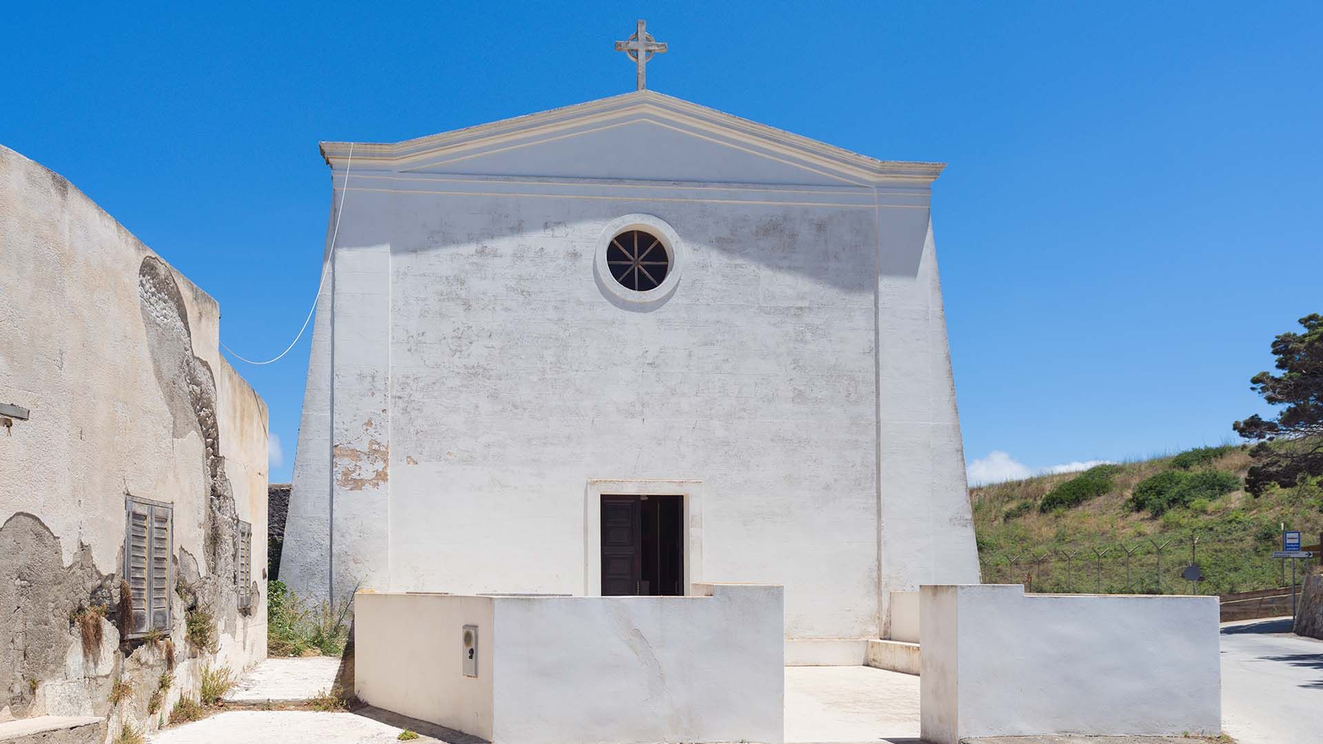 Facciata del Santuario della Madonna della Margana a Pantelleria, edificio semplice intonacato di bianco con timpano triangolare, rosone circolare e croce sulla sommità. Davanti, un piccolo spiazzo recintato da muretti bassi, circondato da un paesaggio ru