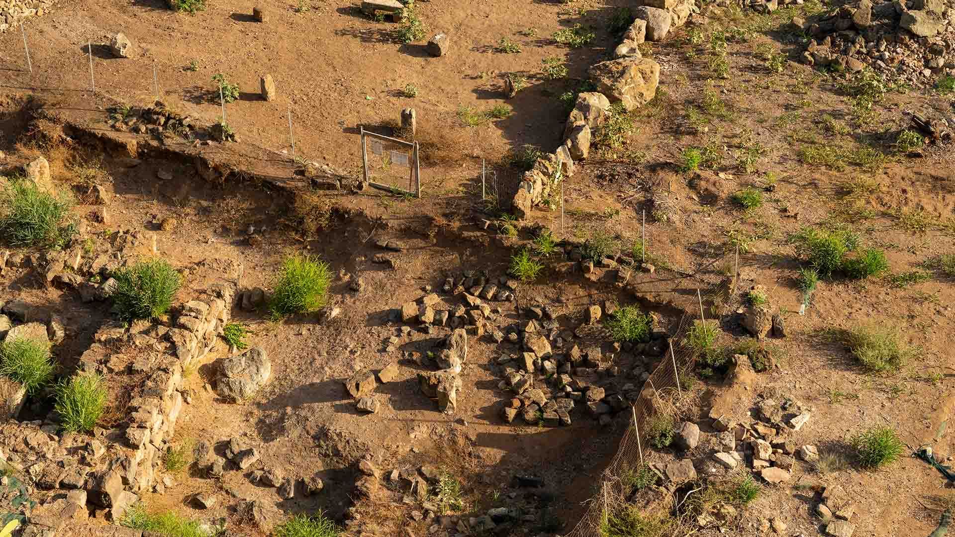 Scavi archeologici all’aperto su un terreno arido, con resti in pietra di antiche strutture sacre e muretti perimetrali. Si notano pietre disposte in forma circolare e rettangolare, e alcune recinzioni metalliche che delimitano l’area protetta.