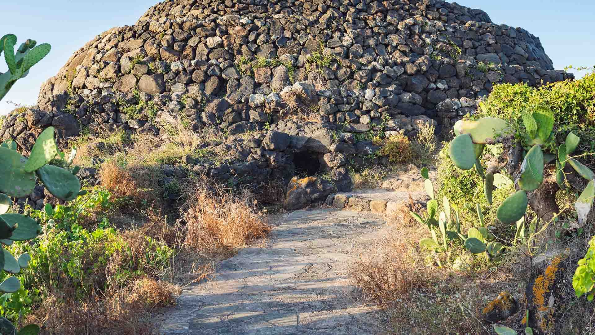 Struttura preistorica in pietra lavica a forma di cupola, composta da blocchi sovrapposti a secco. Un sentiero conduce all’ingresso di una piccola apertura nella base del monumento, circondata da vegetazione spontanea e fichi d’India, sull’isola di Pantel