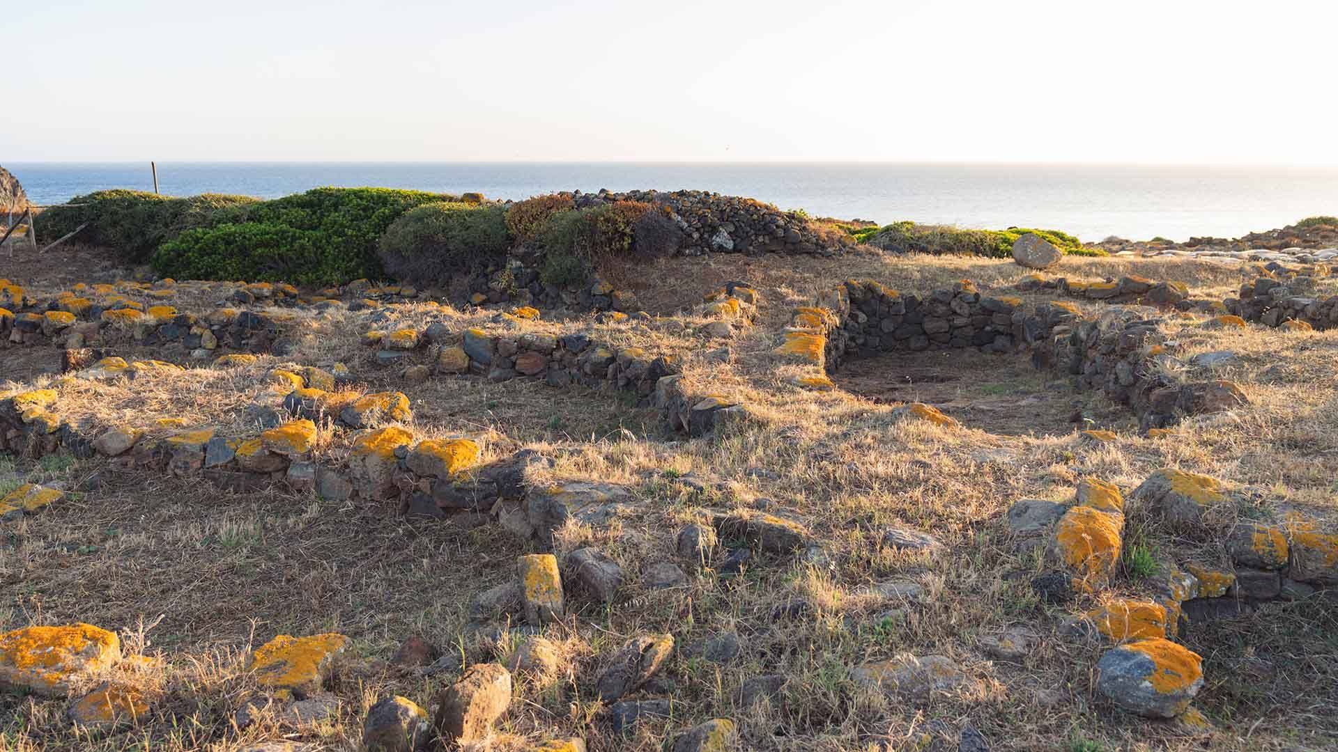 Scavi archeologici in un paesaggio costiero, con muretti a secco in pietra lavica che delineano gli antichi perimetri delle abitazioni preistoriche. Il terreno è arido e coperto di erba secca, con vista sul mare all’orizzonte e cespugli verdi sparsi.