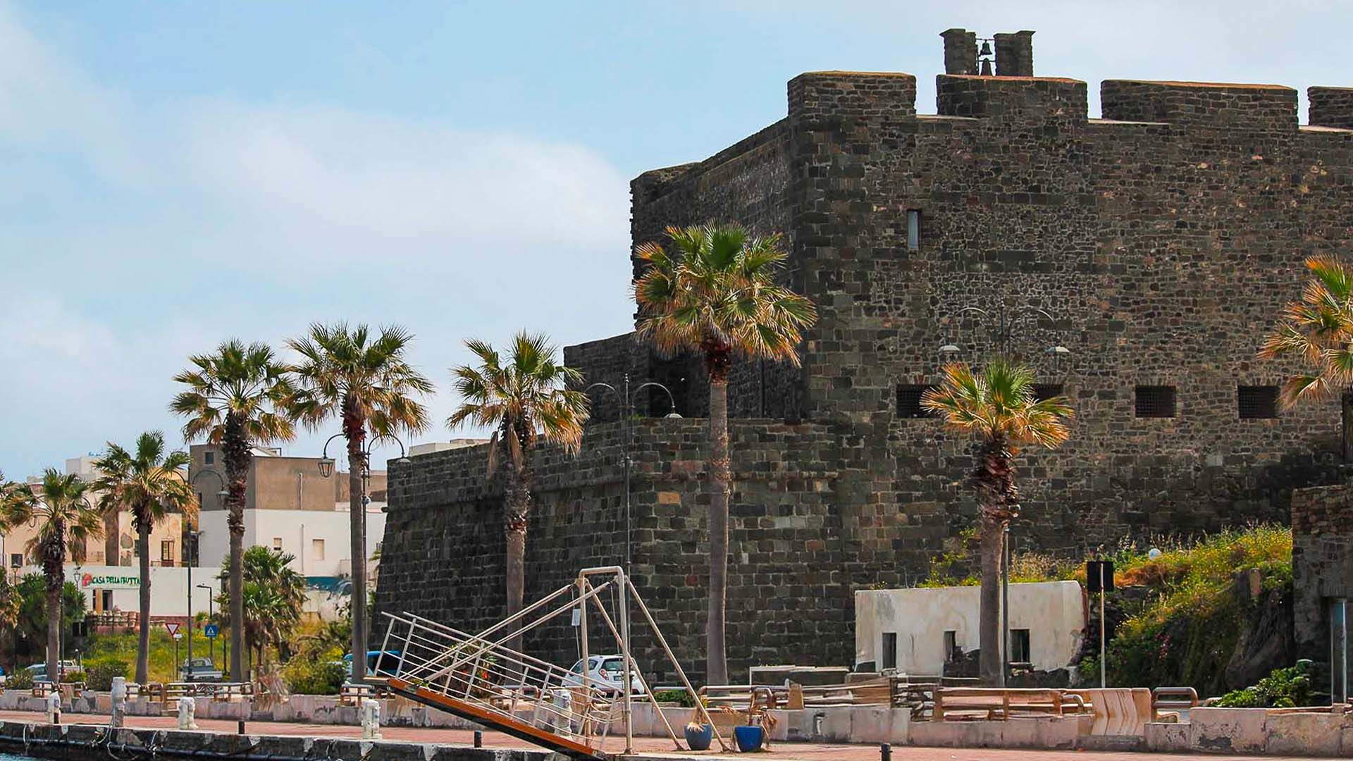 Il Castello di Pantelleria, una fortezza medievale in pietra lavica affacciata sul porto, con palme lungo la banchina.