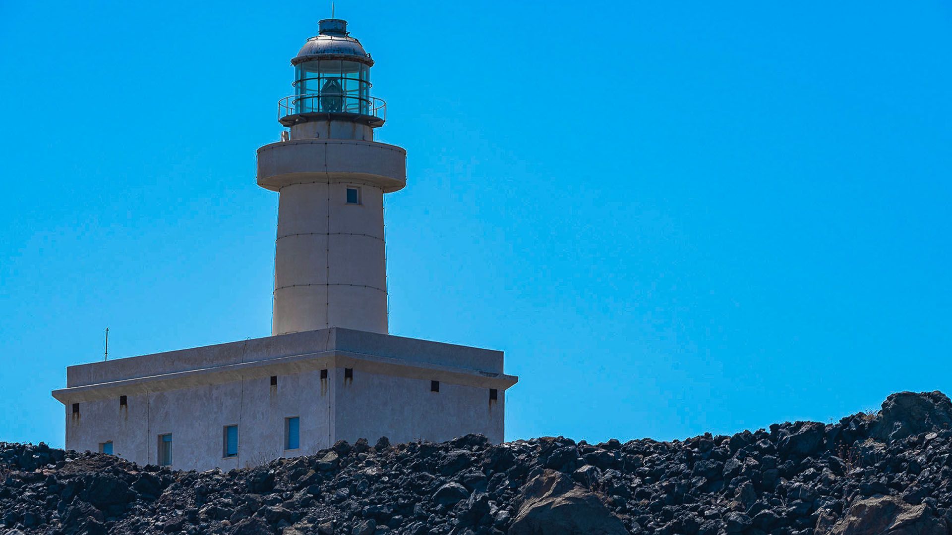 Il Faro di Punta Spadillo a Pantelleria, una torre bianca su rocce laviche con un cielo azzurro intenso.