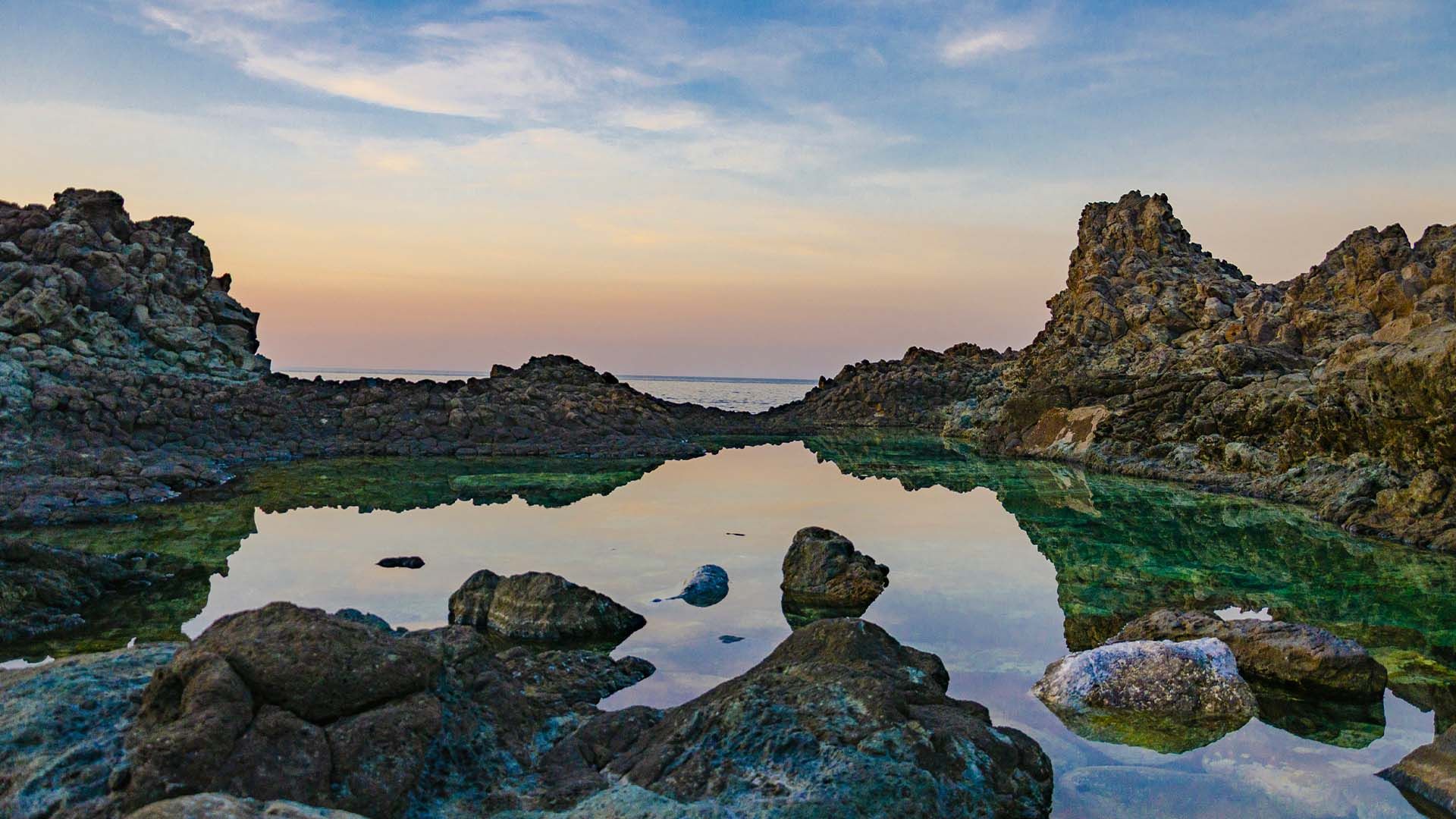 Il Laghetto delle Ondine, una piscina naturale tra le rocce laviche di Pantelleria al crepuscolo con il cielo che si riflette nell'acqua calma.