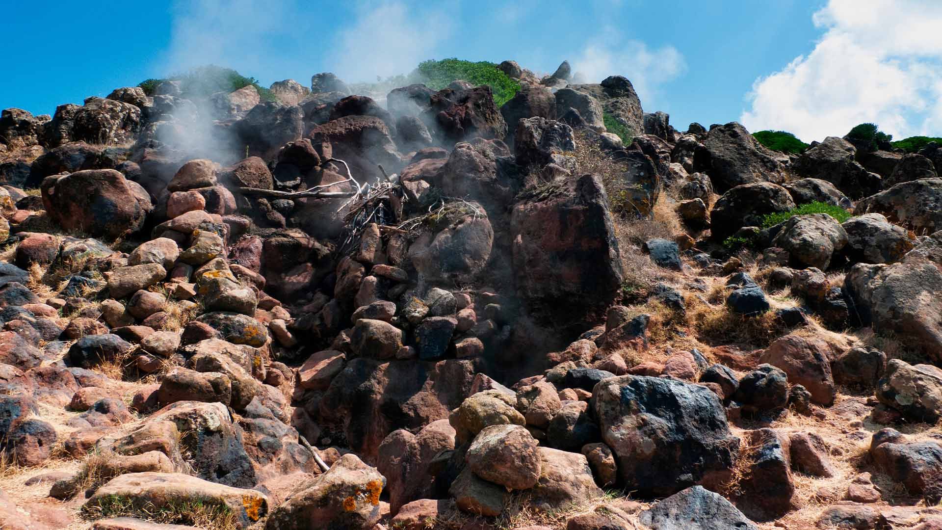 Paesaggio vulcanico di Pantelleria con fumarole attive che rilasciano vapore tra le rocce scure sotto un cielo azzurro.