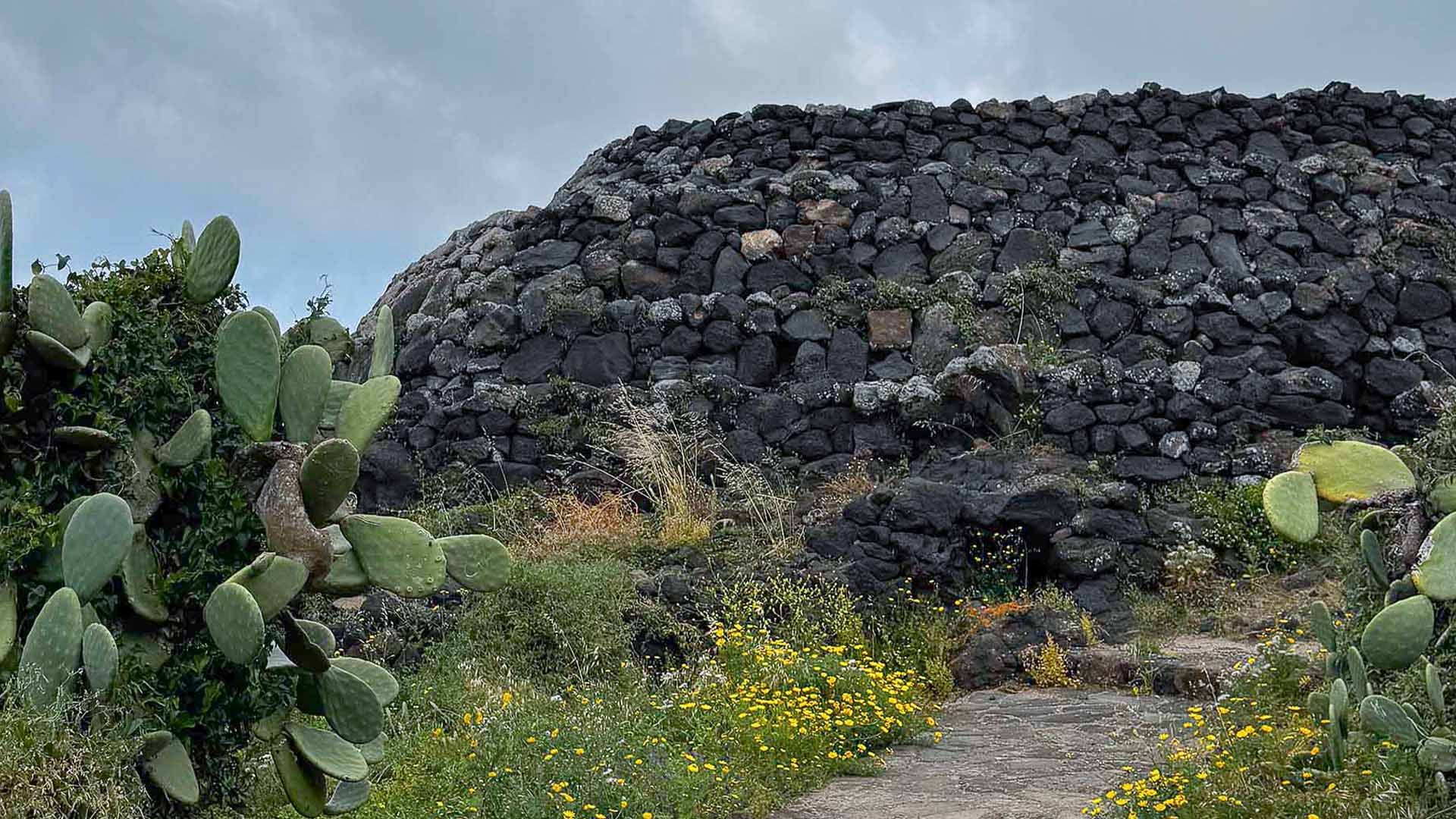 Il Sese del Re, un'antica struttura megalitica funeraria in pietra lavica a Pantelleria, immersa nella vegetazione mediterranea.