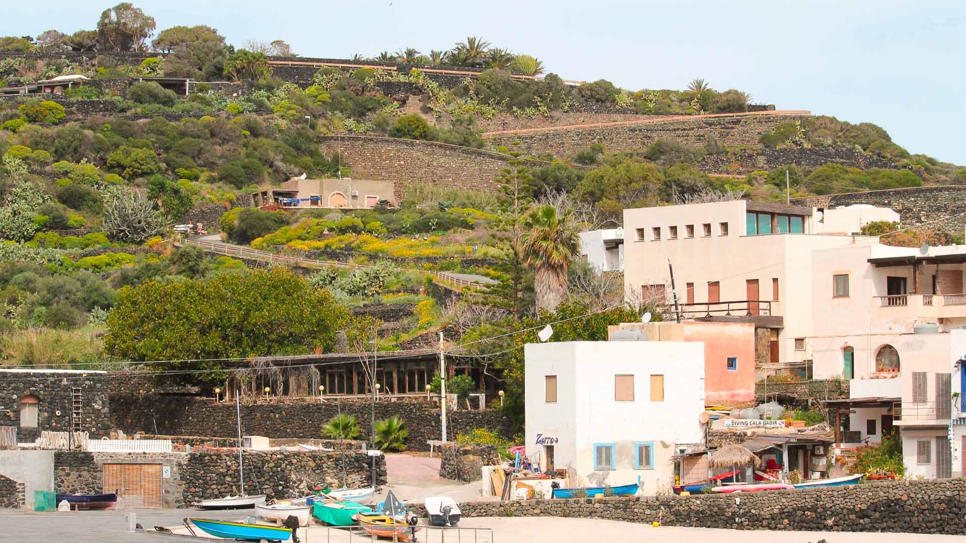 Cala Gadir a Pantelleria con le sue caratteristiche case colorate, la piccola spiaggia, le barche e il paesaggio collinare terrazzato.