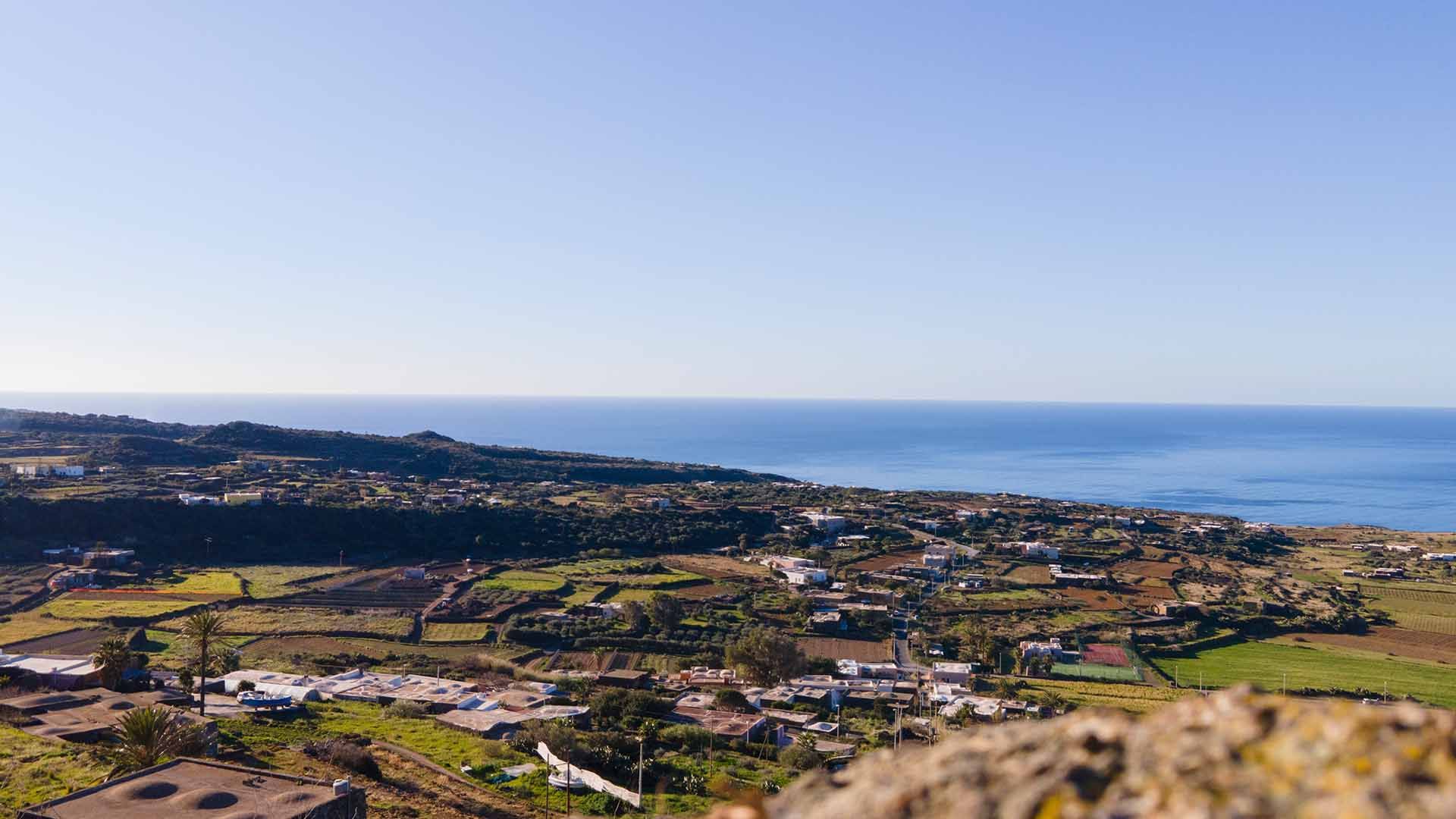 Ampia veduta panoramica di Pantelleria con case sparse, campi coltivati e il mare azzurro sullo sfondo sotto un cielo limpido.