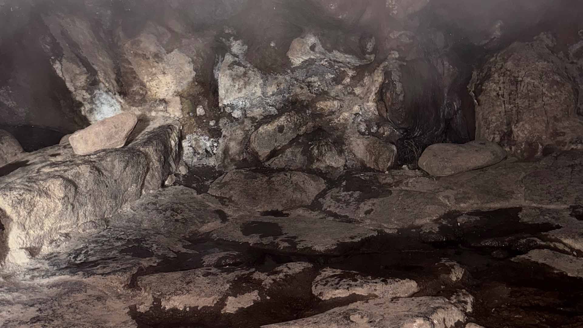 Interno buio di una grotta rocciosa umida con pozza d'acqua scura e pareti irregolari.