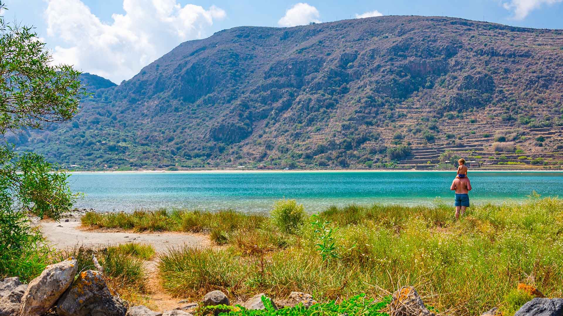 Scena costiera a Pantelleria con un uomo e un bambino vicino a un lago turchese ai piedi di una montagna verdeggiante sotto un cielo sereno.